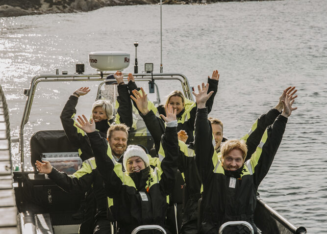 Gruppe med mennesker som står i en båt og hilser mot kamera.